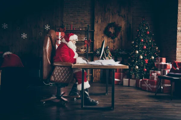 Profiel kant volledige lengte foto gericht grijs haar santa claus zitten tafel werk computer check mail verlanglijstje brieven dragen rood kostuum laarzen in huis binnen met kerst x-mas ornament — Stockfoto