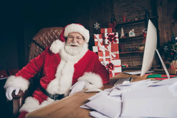 Foto van positieve kerstman zit tafel klaar voor nieuwjaar x-mas voorbereiding cadeau te dragen rood kostuum in huis binnen met x-mas decoratie ornament — Stockfoto