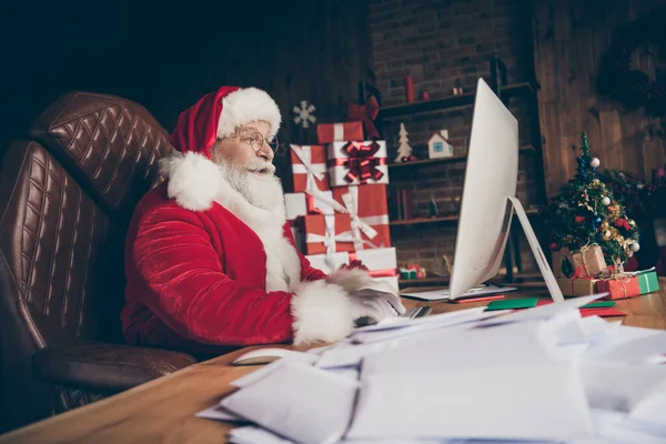 Profiel zijkant foto geschokt grijze baard santa claus zitten tafel met behulp van computer check e-mail onder de indruk verlanglijstje letters dragen rode pet hoofddeksels in huis binnen met x-mas kerst ornament — Stockfoto