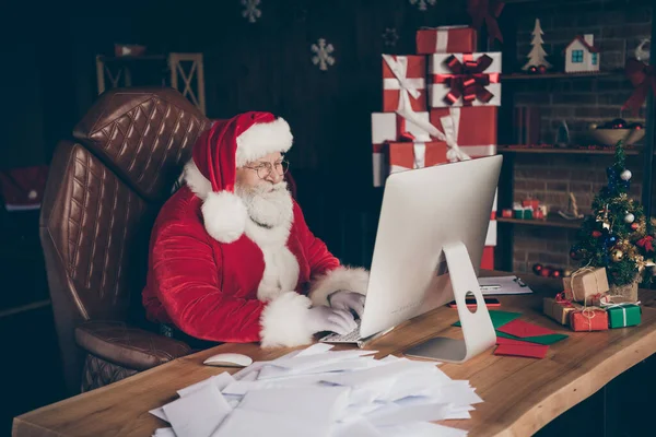 Profilseite Foto von konzentrierten fröhlichen grauen Bart Weihnachtsmann Schreiben Kinder Weihnachten Wunschliste verwenden Computer tragen rote Mütze Kopfbedeckungen sitzen Tisch im Haus drinnen Neujahr Dekoration — Stockfoto