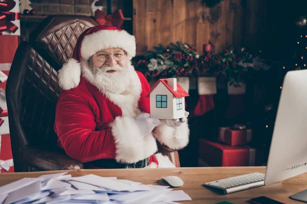 Glückliche Weihnachtseinkäufe. Lustiges weiß-graues Barthaar Weihnachtsmann-Sitztisch hält kleine Versicherungs-Baumütze im Haus drinnen mit Spirituosenatmosphäre adventliches Lametta — Stockfoto