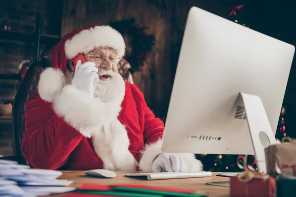 Foto wit grijs baard haar vrolijke hulst santa claus zitten tafel werk computer check e-mail look verlanglijstje brief bel elf smartphone dragen handschoenen pet hoofddeksel in x-mas kerst huis binnen — Stockfoto