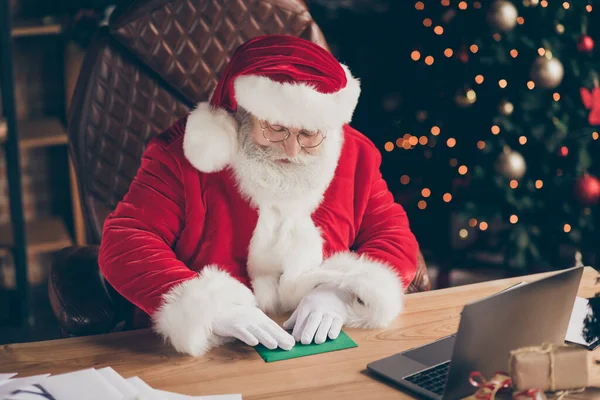 Foto van gerichte hulst vrolijke santa zittafel zegel envelop wilt verzenden brief antwoord op x-mas kerst feest evenement dragen rode pet in huis binnen met nieuwjaar decoratie — Stockfoto