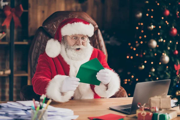 Foto verbaasd grijs haar santa claus zitten tafel in fee noordpool huis lees x-mas kerst kind verlanglijstje onder de indruk cadeau cadeau vraag pet hoofddeksels binnen met nieuwjaar decoratie — Stockfoto