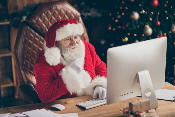 Fee vrolijke hulst santa claus zitten tafel werk computer look scherm lees verlanglijstje brief cadeau e-mail aanraking hand grijze baard in huis x-mas kerst ornament dragen pet hoofddeksel — Stockfoto