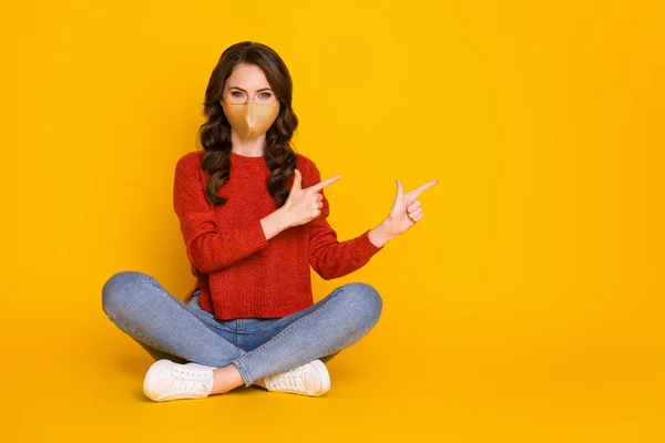 Full längd foto av positiv flicka promotor sitta golv ben korsade punkt pekfinger visar covid nyheter bära denim röd medicinsk mask tröja isolerad ljus glans färg bakgrund — Stockfoto