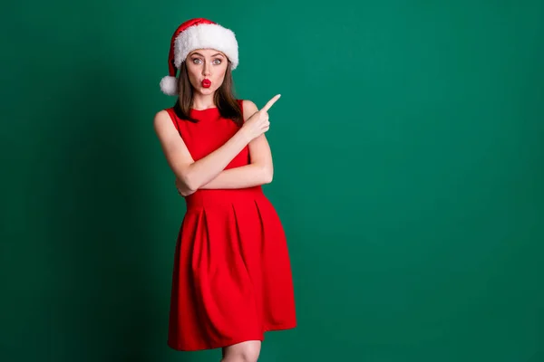 Foto di attraente divertente x-mas negozio assistente signora dito diretto spazio vuoto spettacolo prodotto nuovo anno arredamento prezzi indossare Santa cap rosso mini abito neve ragazza costume isolato colore verde sfondo — Foto Stock