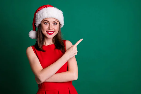 Photo of attractive x-mas shop assistant lady meet customers beaming smile direct finger empty space show novelty wear santa cap red dress snow girl costume isolated green color background