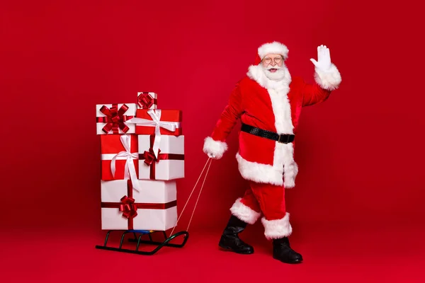 Φωτογραφία πορτρέτο του λευκού γενειοφόρου Santa Claus τραβώντας έλκηθρο με πολλά δώρα κυματίζει το χέρι απομονώνονται σε έντονο κόκκινο χρώμα φόντο — Φωτογραφία Αρχείου