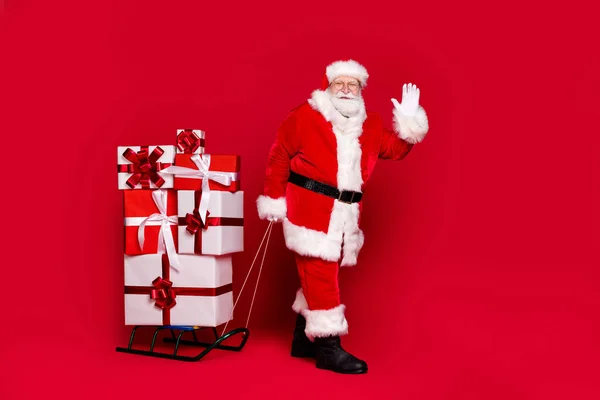 Portrait photo de l'homme barbu blanc funky en costume de Père Noël tirant pile de cadeaux sur traîneau agitant la main isolé sur fond rouge vif — Photo