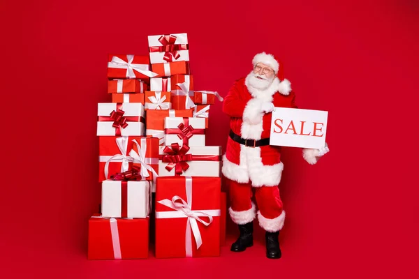 Foto retrato viejo barbudo hombre en traje de Santa Claus celebración de la venta de tarjetas junto a una gran pila de cajas de regalo aislados sobre fondo de color rojo brillante — Foto de Stock