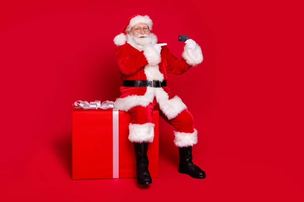Volledige lengte lichaamsgrootte bekijken van zijn hij knappe vet overgewicht vrolijke Kerstman zitten op grote grote giftbox demonstreren plastic kaart geïsoleerde heldere levendige glans levendige rode kleur achtergrond — Stockfoto