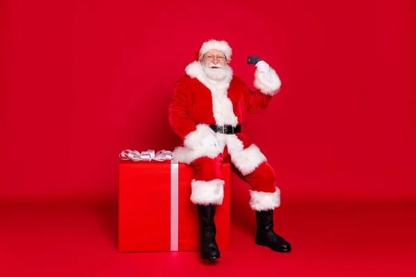 Full length body size άποψη του ο όμορφος λίπος υπέρβαρος Άγιος Βασίλης κάθεται σε μεγάλο μεγάλο giftbox εκμετάλλευση στο χέρι πλαστική κάρτα απομονώνονται φωτεινό έντονο λαμπερό κόκκινο χρώμα φόντο — Φωτογραφία Αρχείου