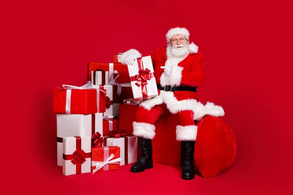 Comprimento total do corpo tamanho vista de seu bonito alegre barbudo Papai Noel desgaste casaco quente sentar na caixa demonstrando entrega giftbox comemorar isolado brilhante brilhante cor vermelha vibrante fundo — Fotografia de Stock