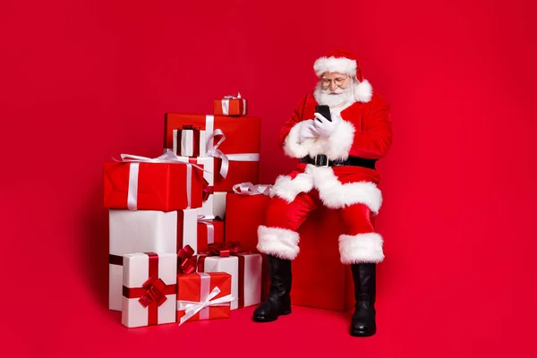 Longitud completa vista del tamaño del cuerpo del traje de Santa Claus con barba guapo enfocado utilizando la tienda de dispositivos de entrega de la pila pila de regalos aislados brillante brillo vivo vibrante color rojo fondo —  Fotos de Stock