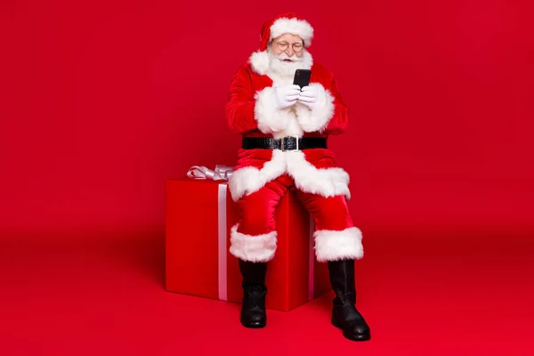 Full length body size view of his he handsome cheerful bearded Santa father wearing warm coat sitting on box use device app delivery celebrate isolated bright vivid shine vibrant red color background — Stock Photo, Image