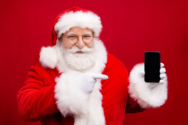 Foto do pensionista vovô barba cinza segurar telefone espaço vazio tela de dedo direto sugerir venda produto x-mas traje luvas brancas óculos casaco headwear isolado cor vermelha fundo — Fotografia de Stock