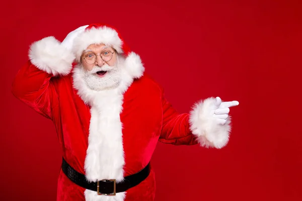 退休老人灰胡子的照片，手头直指空旷的地方，暗示着疯狂的新奇穿着桑塔圣诞服装皮带手套眼镜头戴孤立的红色背景 — 图库照片
