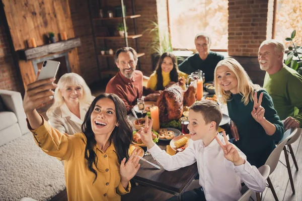 Photo of full family gathering eight people woman mother hold telephone make shoot selfie little kid grandma show v-sign dinner big table turkey generation home evening living room indoors