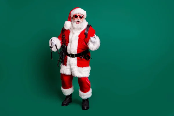 Взгляд в полный рост на его привлекательного веселого сумасшедшего холодного бородатого веселого Деда Мороза, держащего в руках плетеные инструменты, показывающие знак рога, веселящегося на изолированном зеленом цветовом фоне — стоковое фото