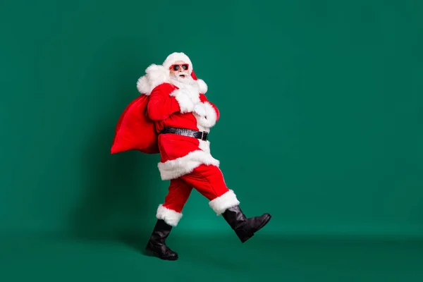 外公白胡子背着背包礼物走着的全长相貌照片，孩子们穿着红色圣诞老人服装手套太阳镜头饰孤立的绿色背景 — 图库照片