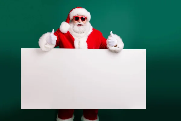 Photo of retired stylish grandfather grey hair beard hold white board thumb-up approve promo wear red santa x-mas costume coat gloves sunglass headwear isolated green color background — Stock Photo, Image