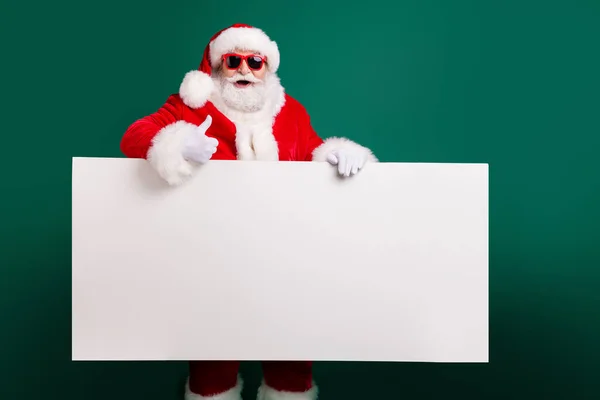 Photo of retired stylish grandfather grey hair beard hold white board thumb-up assure chosen object wear red santa x-mas costume coat gloves sunglass headwear isolated green color background — Stock Photo, Image