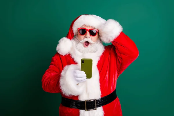 Foto van gepensioneerde stijlvolle grootvader grijs haar baard houden telefoon verbaasd open mond hoofd dragen rode santa x-mas kostuum jas handschoenen zonneglas kap geïsoleerde groene kleur achtergrond — Stockfoto