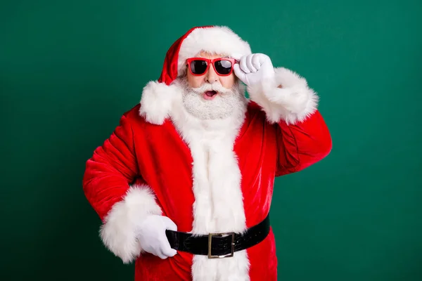 Ez hihetetlen. Fénykép a nyugdíjas nagypapa fehér szakáll nyitott száj tartsa szemüveg nagy hóember olvad viselni piros x-mas mikulás jelmez kabát kesztyű napszemüveg fejfedő elszigetelt zöld színű háttér — Stock Fotó