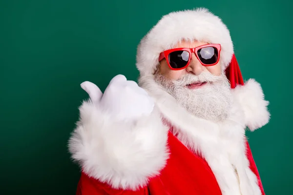 Primo piano headshot foto del nonno in pensione barba bianca pollice diretto spazio vuoto newyear sconto indossare rosso Santa x-mas costume cappotto guanti occhiali da sole copricapi isolato colore verde sfondo — Foto Stock