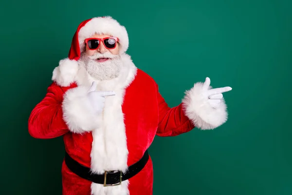 Foto van gepensioneerde opa grijze baard glimlach directe vinger lege ruimte bieden nieuwjaar decoraties dragen rode kerstman x-mas kostuum jas handschoenen zonneglas riem hoofddeksel geïsoleerde groene kleur achtergrond — Stockfoto