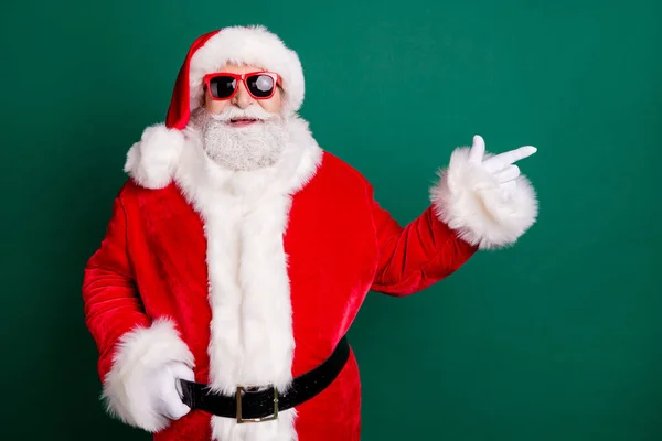养老金领取者祖父白胡子笑着直接手指空空荡荡的地方提出产品穿红色桑塔圣诞服装手套太阳镜带头饰孤立绿色背景 — 图库照片
