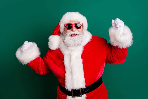 Фото пенсионера дедушки седой бороды улыбаясь поднять кулаки весело праздник дискотека носить красный Санта-мас костюма пальто перчатки солнцезащитные очки головной убор изолированный зеленый цвет фона — стоковое фото