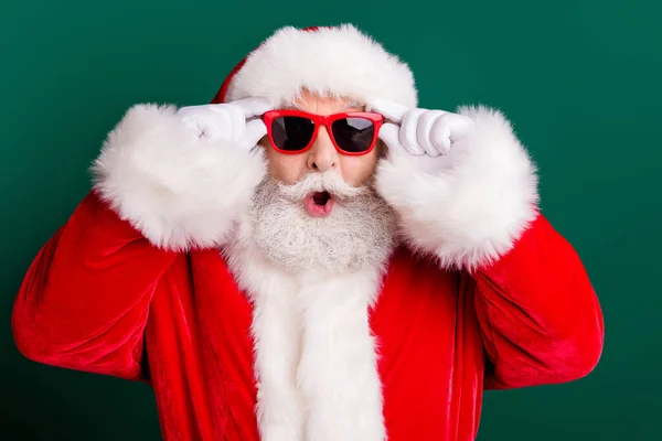Closeup portrait photo of retired old man white beard open mouth hold eyewear shocked see unbelievable big sleigh wear red santa costume sunglass headwear isolated green color background — Stock Photo, Image
