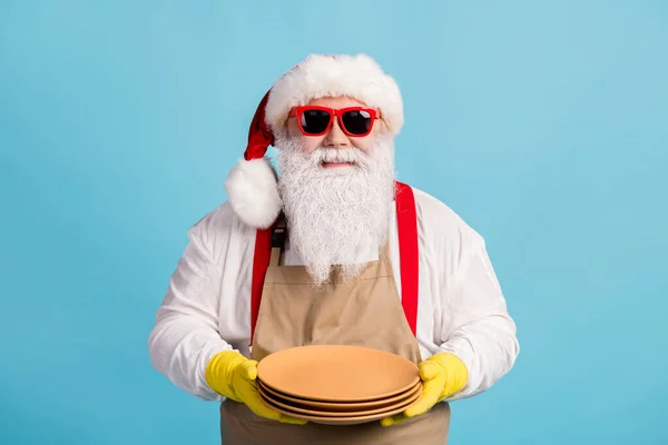 Photo of pensioner old man grey beard hold clean dishes restaurant service waiter half-time wear santa x-mas costume apron rubber glove suspender sunglass cap isolated blue color background