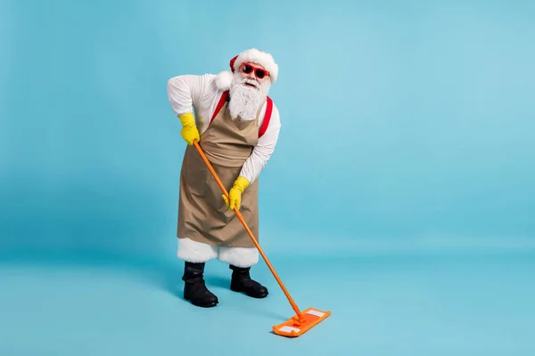 Pleine taille photo du vieil homme à la retraite barbe grise tenir balai lavage étage public concierge porter Santa x-mas costume tablier en caoutchouc gant bretelles sunglass cap isolé fond de couleur bleue — Photo