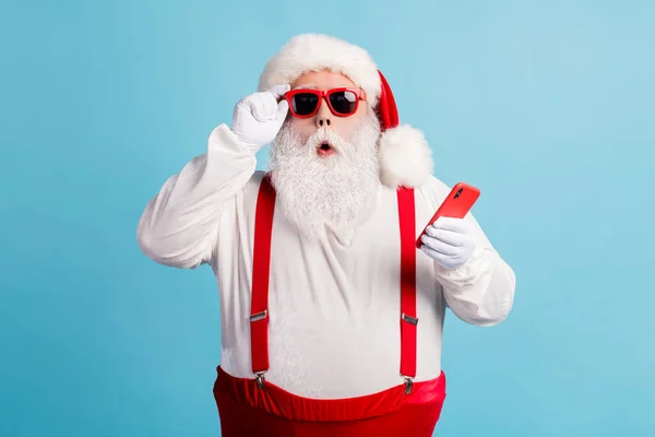 Foto di pensionato nonno grigio barba tenere telefono leggere fake news Natale annullato usura Santa x-mas rosso costume bretelle occhiali da sole guanti bianchi cappello isolato colore blu sfondo — Foto Stock