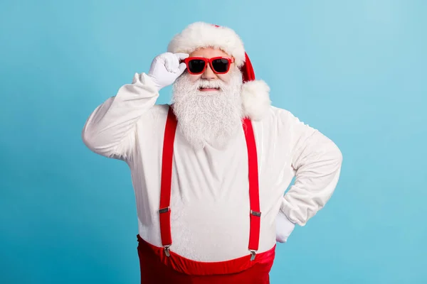 Foto van gepensioneerde oude man grijze baard houd specs hand heup zelfverzekerd voor te bereiden check slee dragen santa kostuum rode bretels brillen witte handschoenen hoofddeksel geïsoleerde blauwe kleur achtergrond — Stockfoto