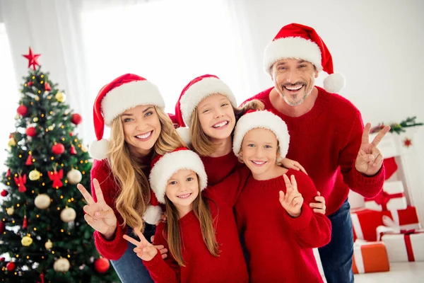 Foto porträtt av rolig vänlig familj bär varma röda festliga tröjor och huvudbonader förbereder sig för jul kväll nära xmas träd dekorationer visar v-tecken med två fingrar — Stockfoto