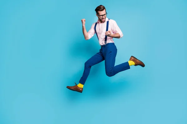 Teljes méretű profil fotó jóképű férfi ugrás magasra futó verseny emelje öklöt első helyen győztes verseny maraton kopás szemüveg ing nadrágtartó nadrág csizma elszigetelt kék háttér — Stock Fotó