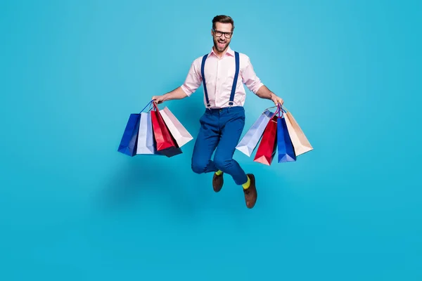 Full length photo of handsome business man carry bags jump high up buy holiday stuff shopping center store mall wear specs shirt suspenders pants shoes isolated blue color background — Zdjęcie stockowe