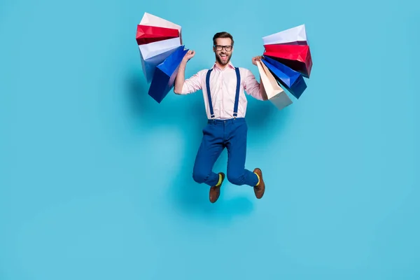 Foto de tamaño completo de hombre de negocios guapo llevar bolsas saltar alto comprar cosas de vacaciones centro comercial buen humor desgaste especificaciones camisa tirantes pantalones botas aislado color azul fondo —  Fotos de Stock