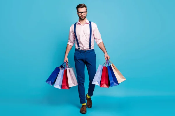 Πλήρης φωτογραφία μεγέθους του όμορφος επιχειρηματίας επιτυχημένος εργαζόμενος μεταφέρουν τσάντες αγοράζουν ρούχα διακοπές εμπορικό κέντρο φορούν specs πουκάμισο τιράντες παντελόνι μπότες κάλτσες απομονωμένο παστέλ μπλε χρώμα φόντο — Φωτογραφία Αρχείου