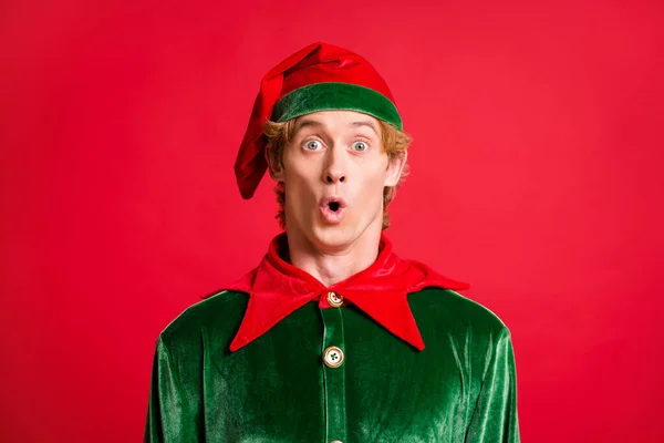 Portrait of astonished elf guy look in camera unbelievable news hear wear green costume hat isolated on red color background