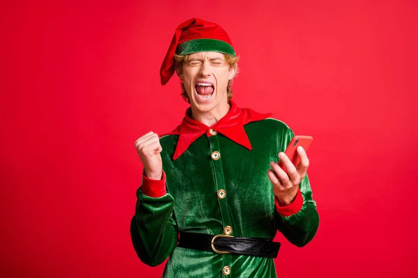 Фото сумасшедшего восторженного X-mas эльф парень держать мобильный телефон поднять кулаки носить зеленый костюм изолированы на красном ярком фоне цвета — стоковое фото