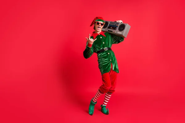 Çılgın elf 'in müzik kutusunun tam boy fotoğrafı. Noel şarkıları boynuzlu sembolü gösteriyor. Kırmızı arka planda izole edilmiş yeşil kostüm giyiyor. — Stok fotoğraf