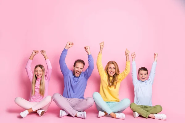 Full size photo of ecstatic dad mom and small girl boy sit with crossed legs raise fists isolated on pastel color background