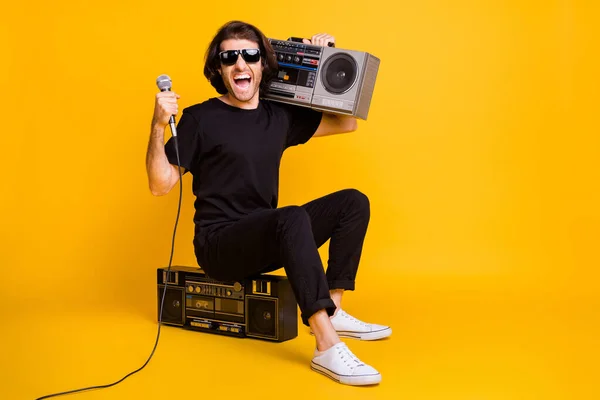 Genç adamın tam boy fotoğrafı mikrofon açık omuz kasetçalar otur siyah t-shirt pantolon giy güneş gözlüğü izole sarı arka plan — Stok fotoğraf