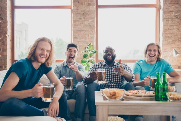 Retrato de locos cuatro amigos de la gente sentarse comodidad sofá reloj broma comedia serie celebrar cerveza pinta taza dedo riendo tienen patatas fritas en casa en el interior — Foto de Stock