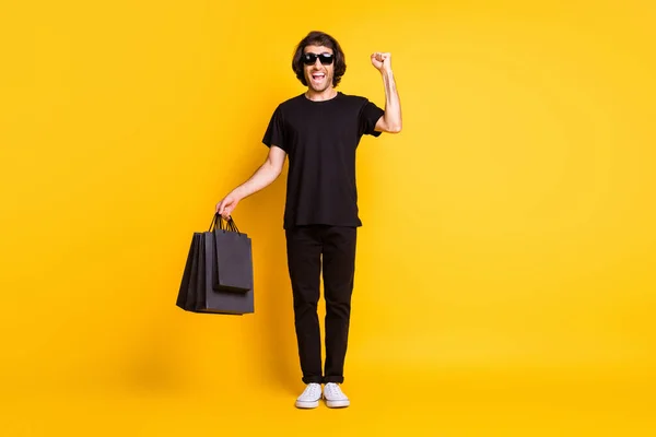 In voller Länge Foto von jungen Mann stehen heben Arm Faust halten Taschen tragen schwarzes T-Shirt Hose weiße Turnschuhe Brille isoliert gelbe Farbe Hintergrund — Stockfoto